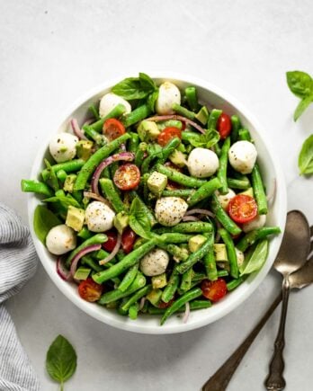 White bowl with green bean salad and mozzarella balls next to spoon and fork.