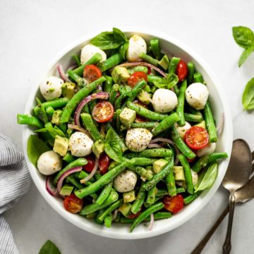 White bowl with green bean salad and mozzarella balls next to spoon and fork.