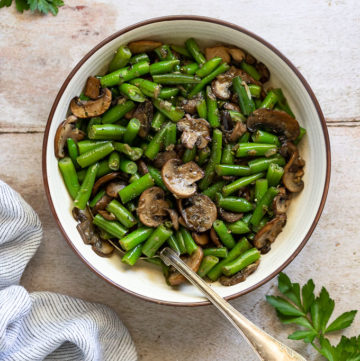 Serving bowl of green beans and mushrooms with spoon next to parsley.