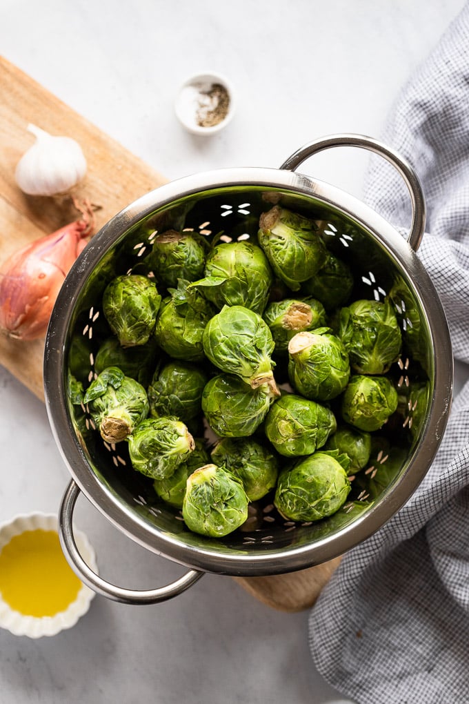 Bowl of Brussels sprouts next to shallot, garlic, and olive oil.