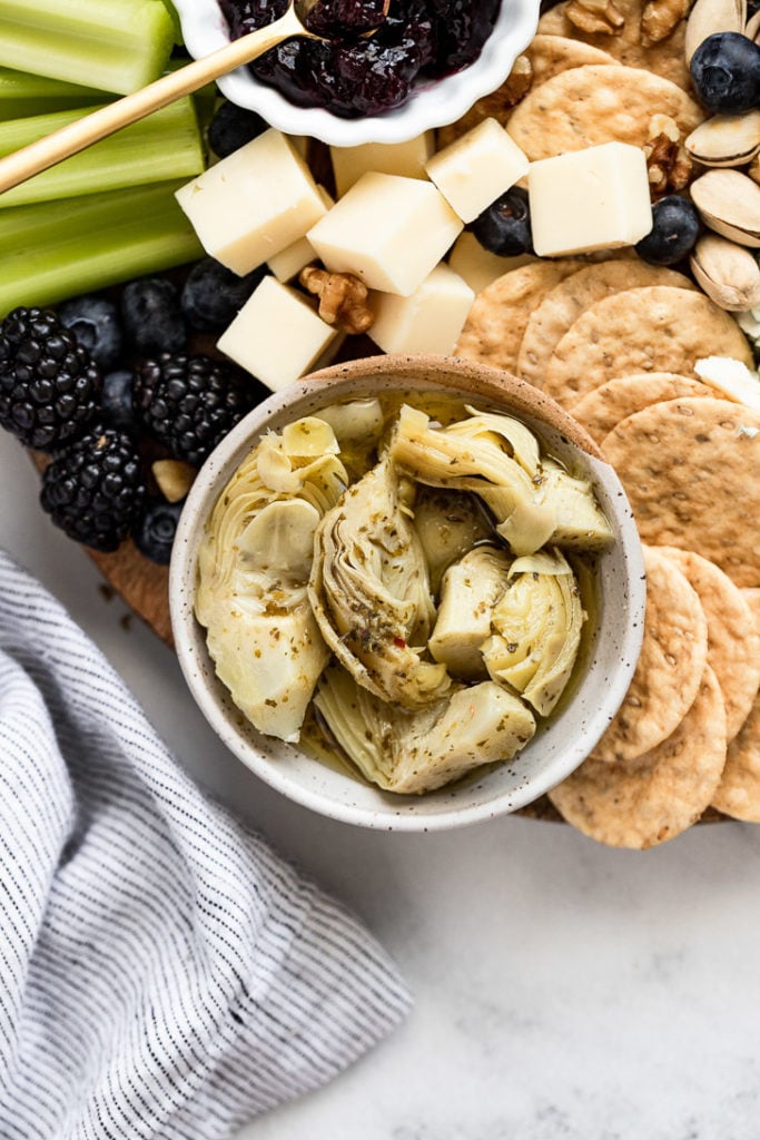 Marinated artichokes in small bowl on board next to cheese, crackers,and berries.