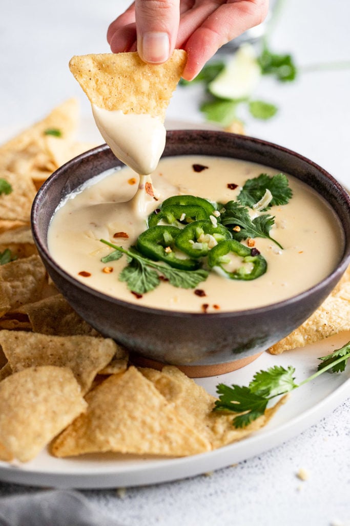 Chip dipping into bowl of queso garnished with jalapeno next to tortilla chips.