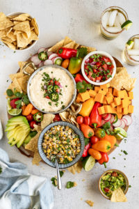 Board with three dips, chips, peppers, and cheese.