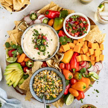 Board with three dips, chips, peppers, and cheese.