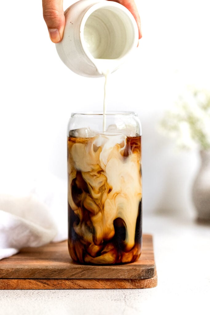 Cream pouring into glass of iced coffee on wood tray.