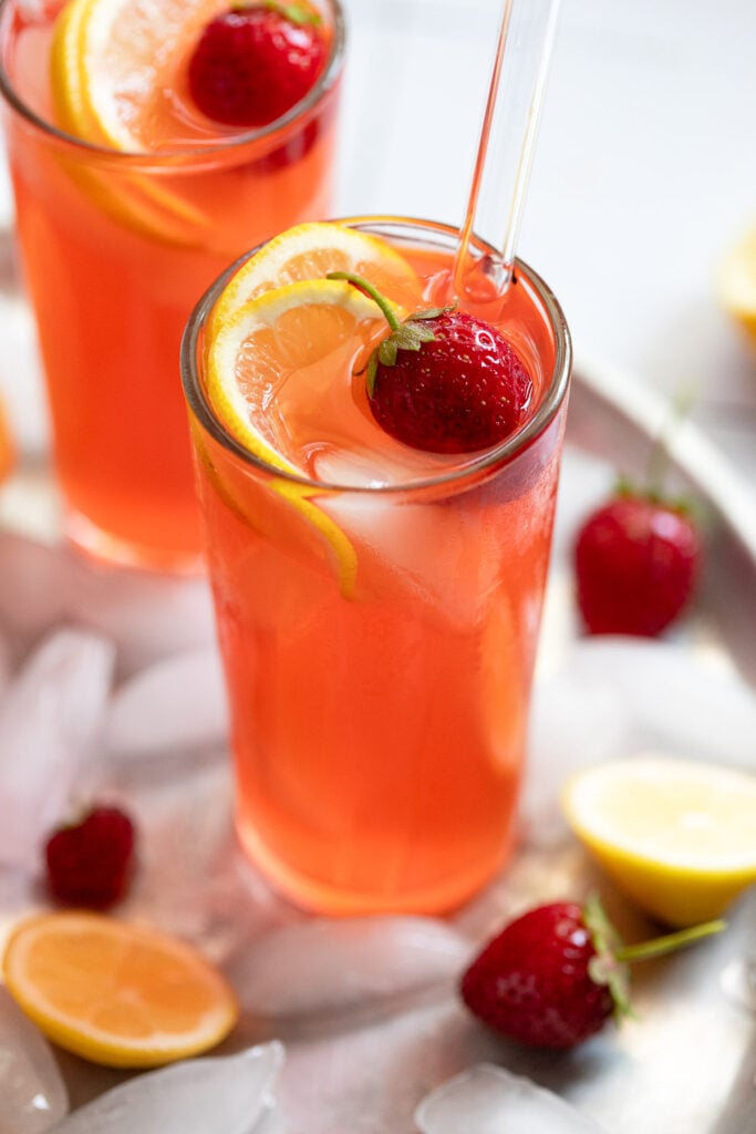 Two glasses of strawberry lemonade on tray with ice, lemons, and strawberries.