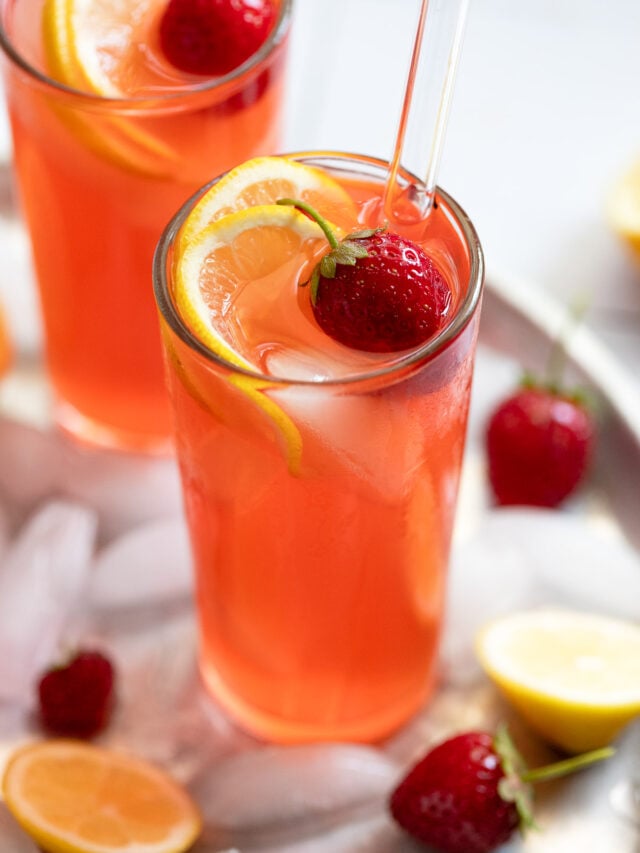 Two glasses of strawberry lemonade on tray with ice, lemons, and strawberries.