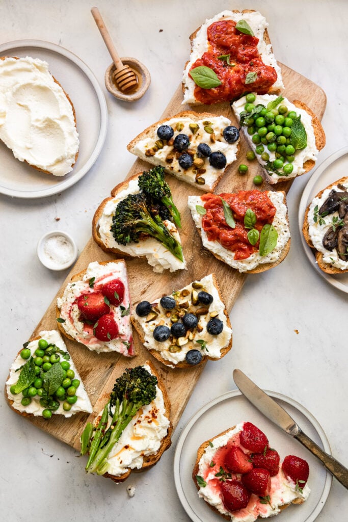 Ricotta toasts with various toppings cut in half across board and plates.