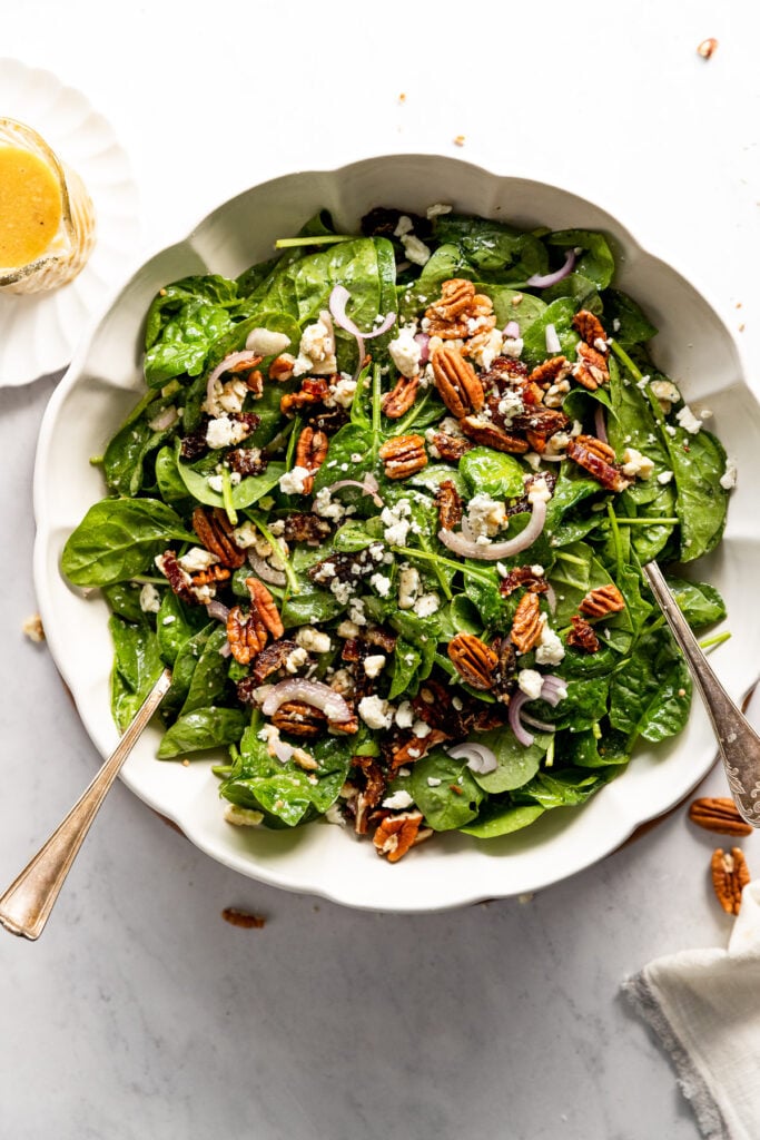 Bowl of spinach salad with serving spoons next to dressing.