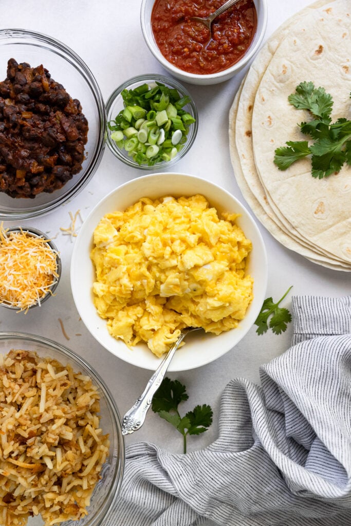 Scrambled eggs in bowl next to spicy beans, hash browns, tortillas, salsa, and cheese.