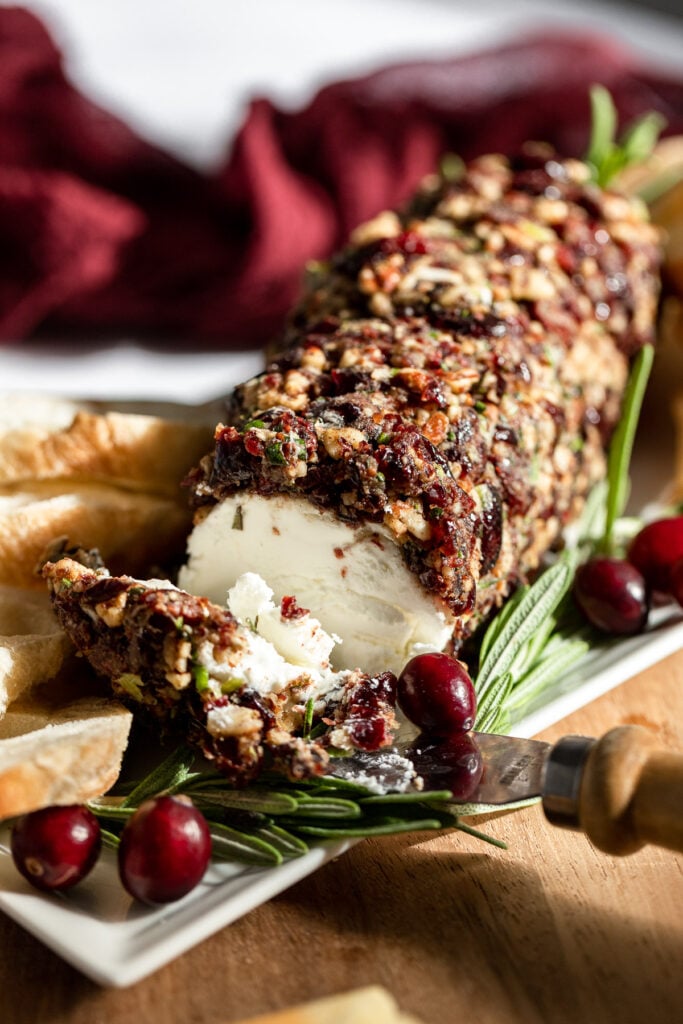 Cranberry goat cheese log with slice out next to bread and rosemary.