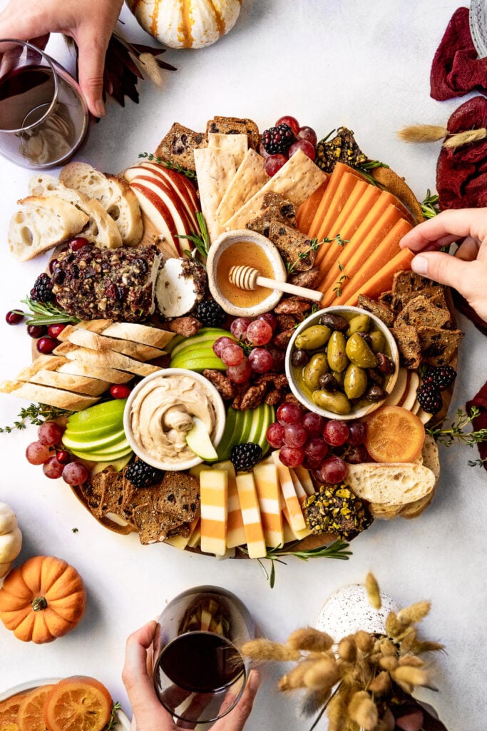 Hands holding wine around fall snack board and grabbing cheese.