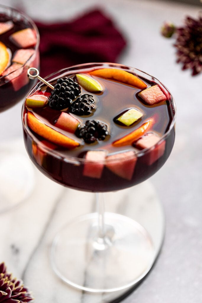Coupe glass with sangria topped with blackberries, apple, and orange slices.