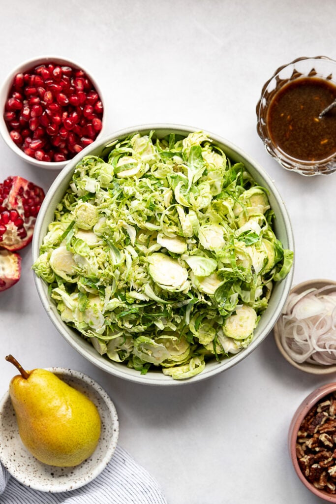 Shredded brussels in bowl next to pomegranate, dressing, shallot, pecans, and pear.