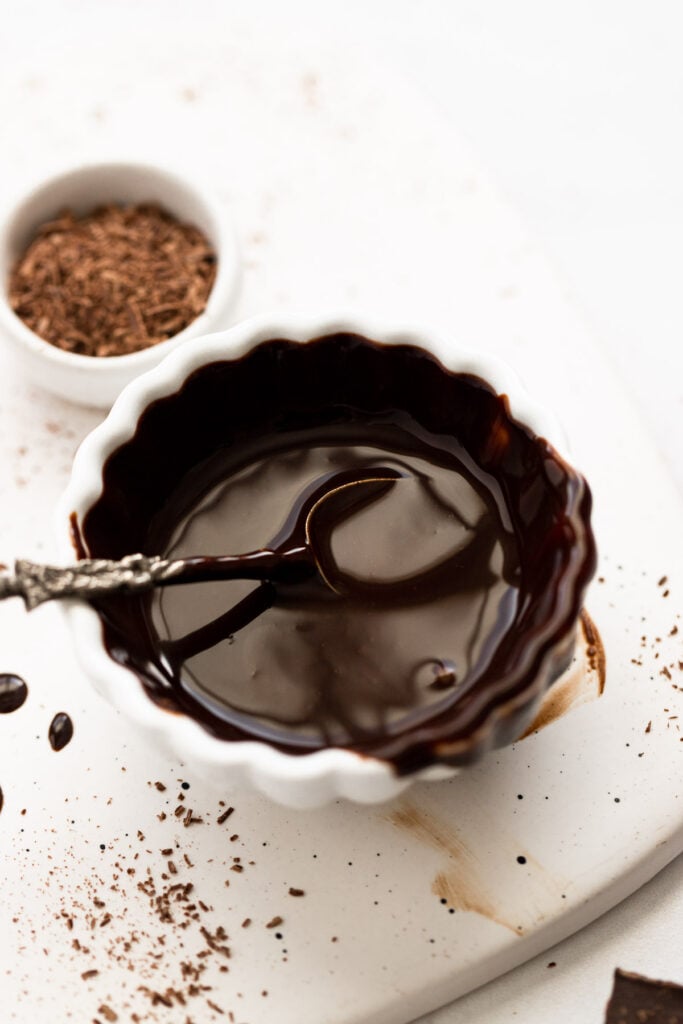 Chocolate syrup in bowl with spoon next to chocolate shavings.