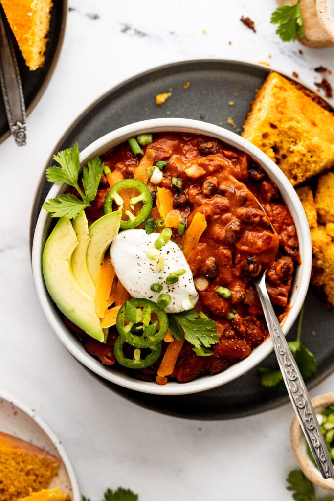 Bowl of vegetarian chili topped with sour cream, avocado, jalapeno next to corn bread with spoon in it.