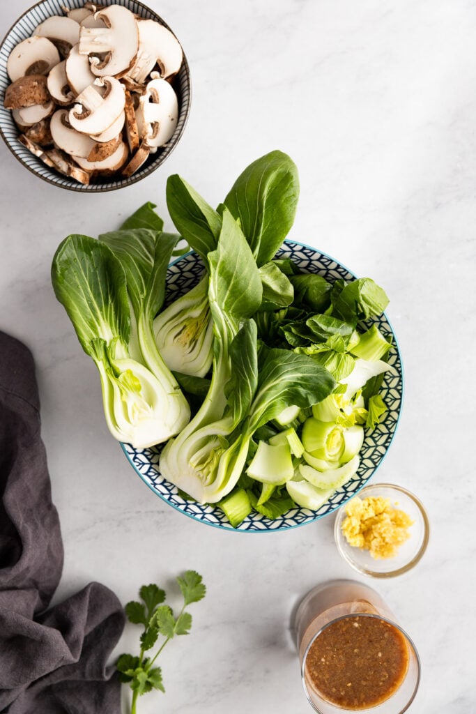 Bowl of bok choy next to bowl of sliced mushrooms, grated ginger, and stir fry sauce.