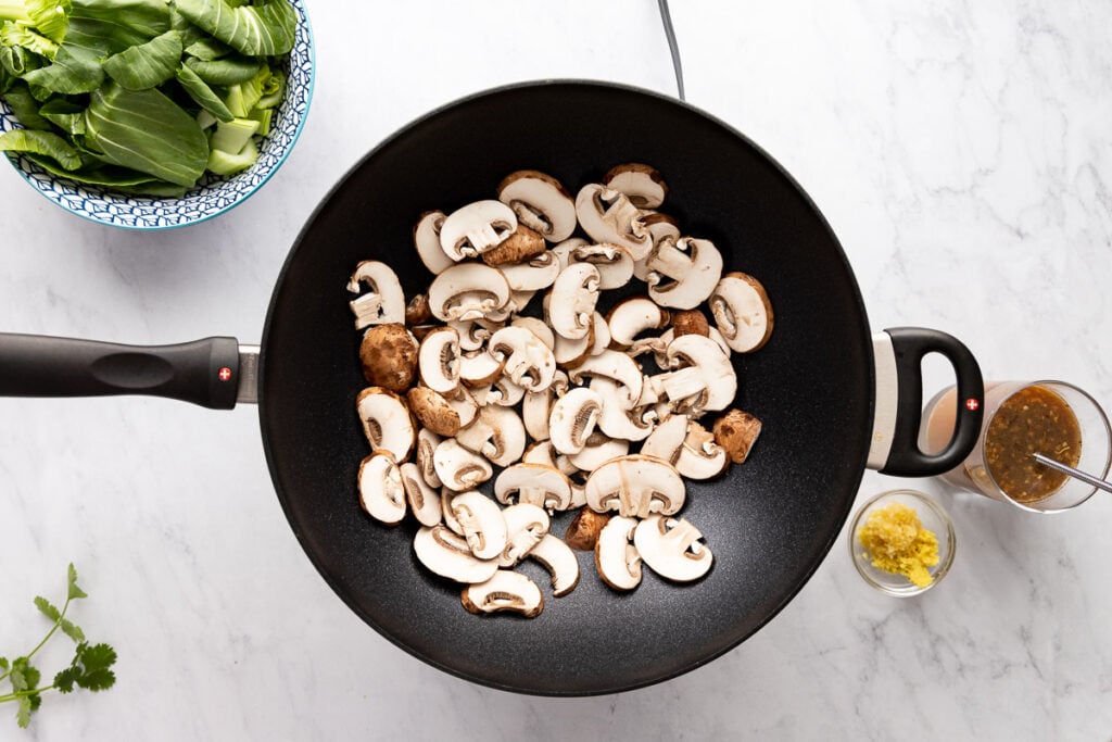 Sliced mushrooms in wok with bowl of bok choy and sauce on side.
