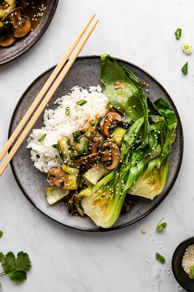 Plate of bok choy stir fry with mushrooms with rice and chopsticks next to it.