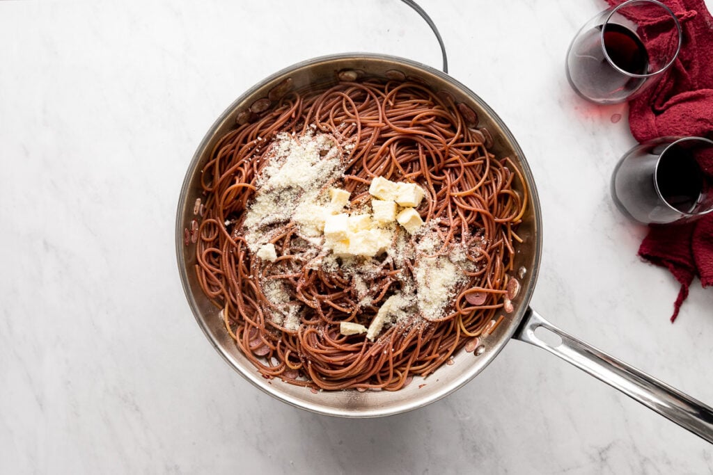 Skillet with red wine noodles topped with butter cubes and cheese.