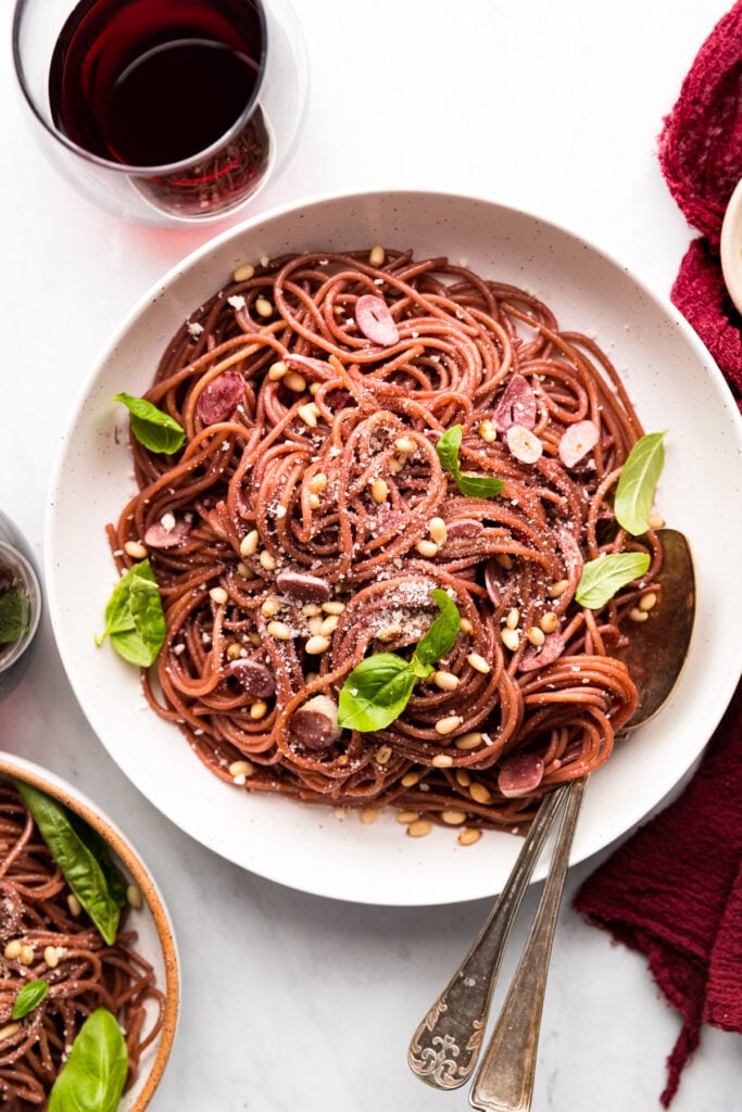 Bowl of red wine pasta with basil leaves, pine nuts, and serving spoon inside.