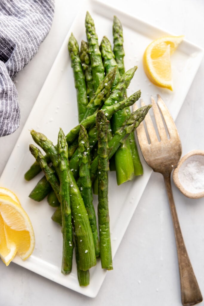 Asparagus spears on white tray with serving fork and lemon wedges.