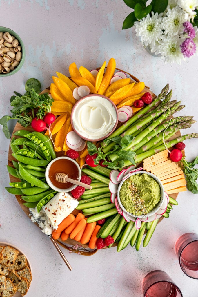 Round board with asparagus, bell peppers, three bowls, cucumber, cheese slices, radishes.