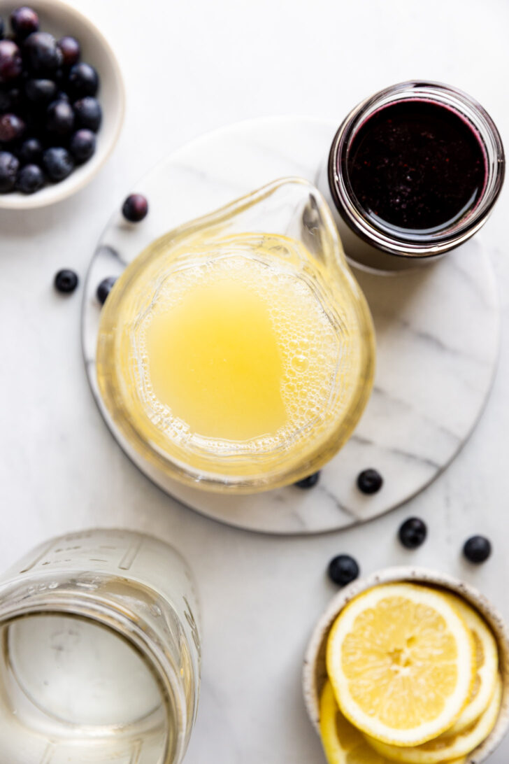Pitcher of lemon juice next to blueberry syrup and lemon slices.