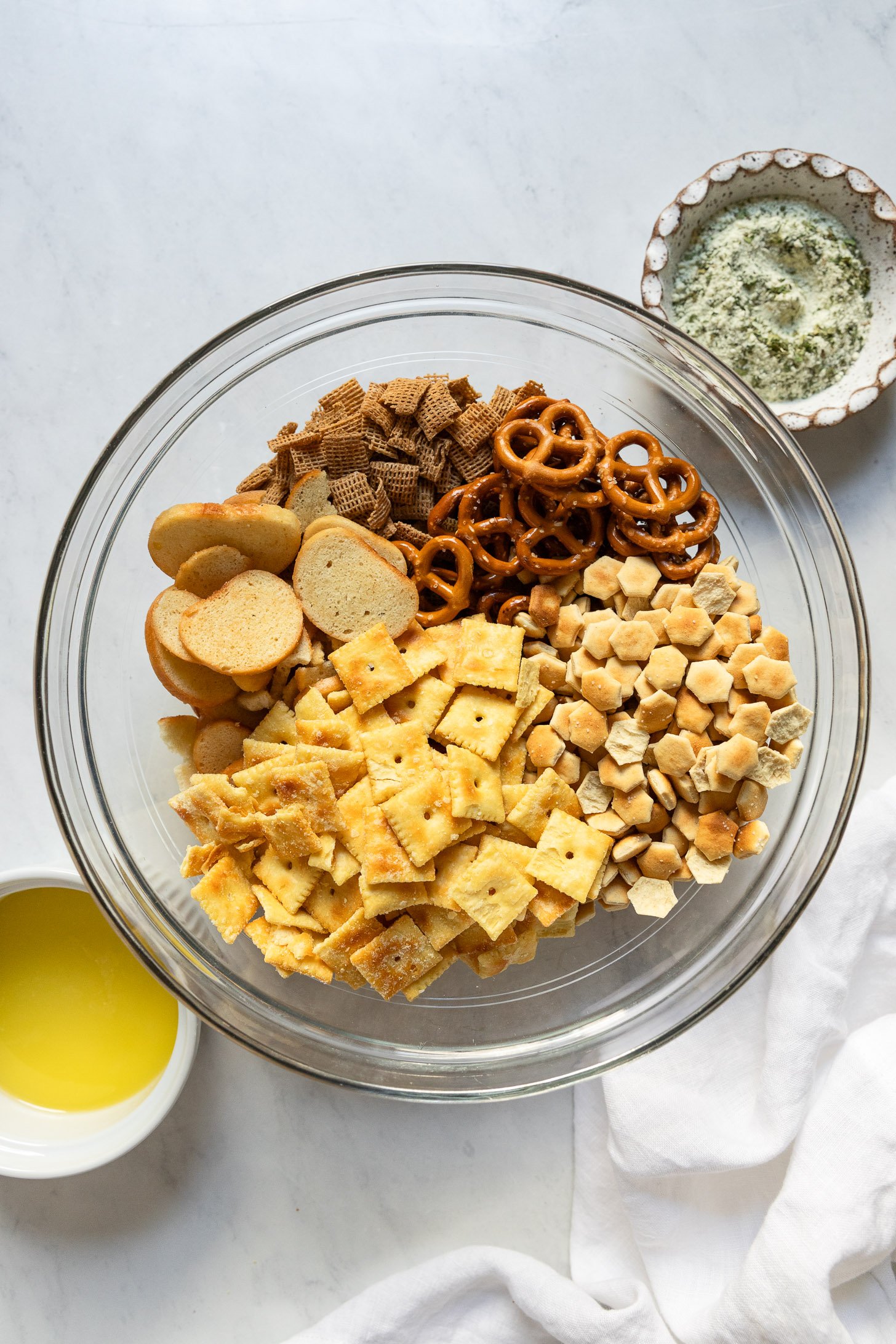 Bowl of bagel chips, cheese crackers, oyster crackers, pretzels, and wheat cereal next to bowls of ranch seasoning and melted butter.
