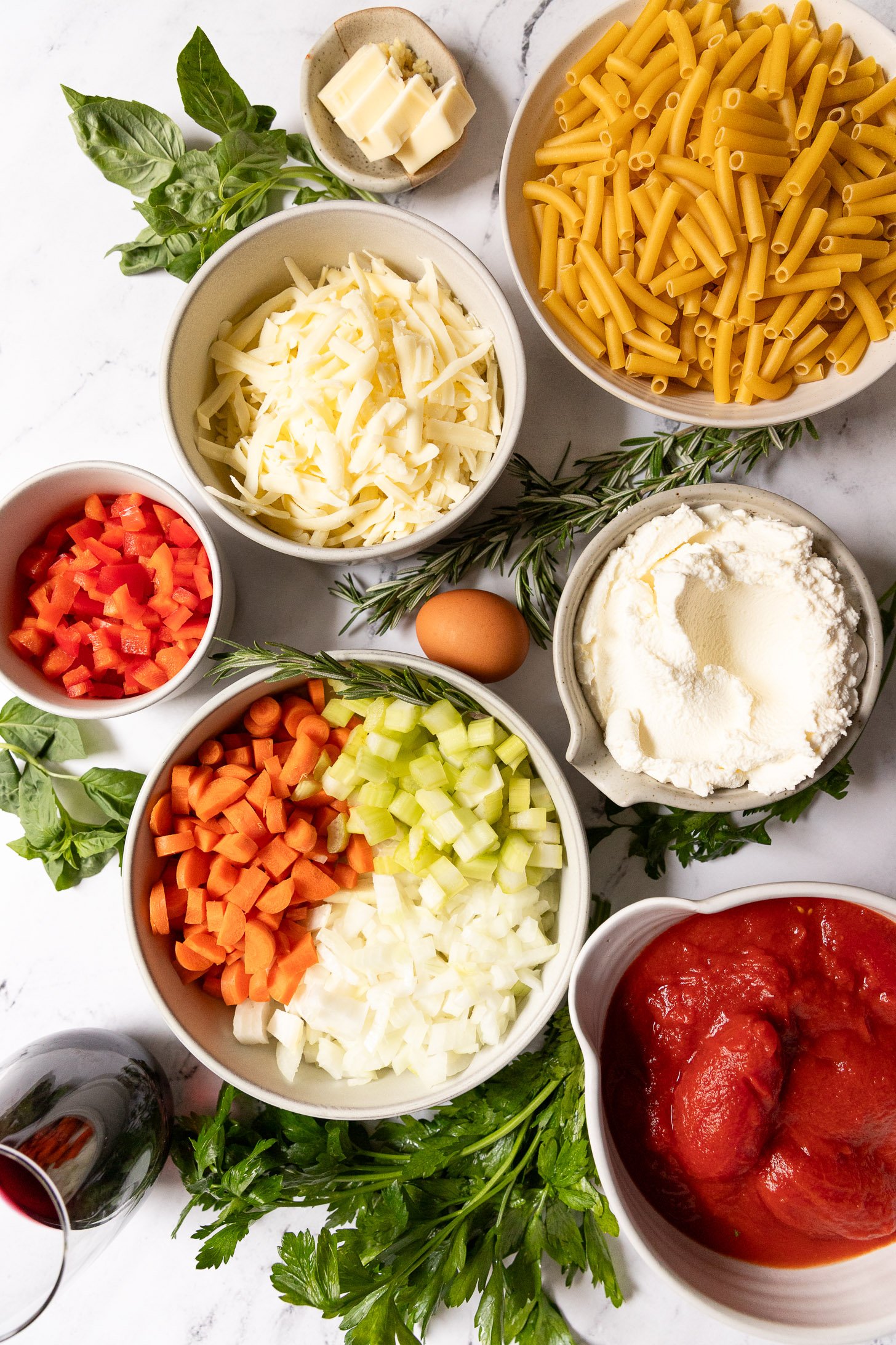 Bowls of ziti ingredients surrounded by sprigs of fresh herbs.