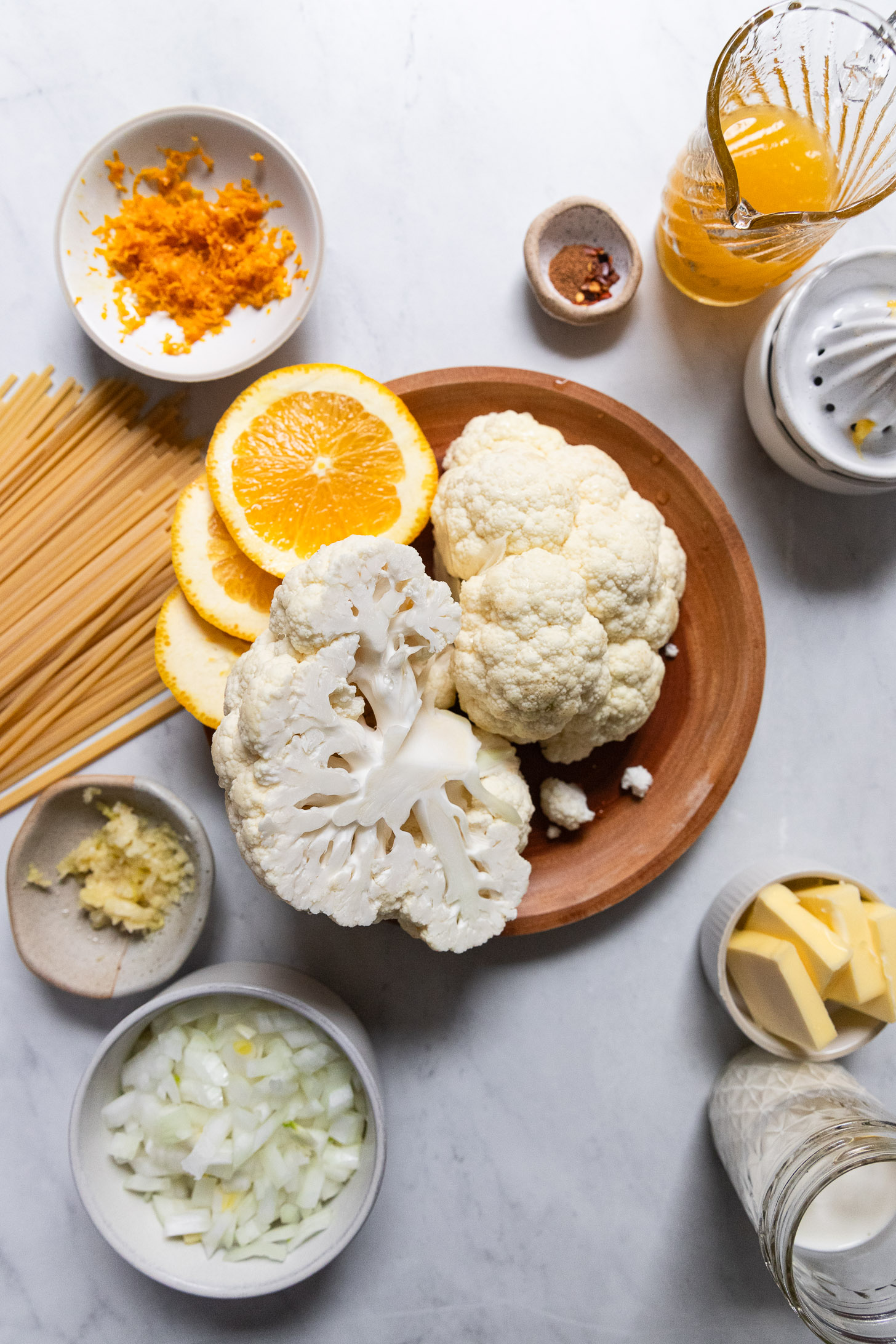 Cauliflower head cut in half with orange slices next to dried pasta, orange zest, onion, and jars of liquids.