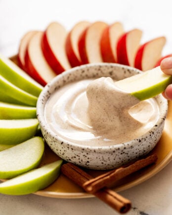 Apple slice dipping into yogurt dip surrounded by plate of apple slices.