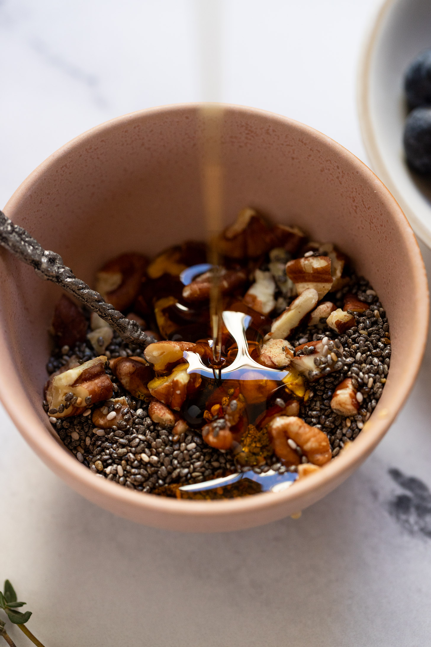 Maple syrup drizzling into bowl of chopped pecans and chia seeds.