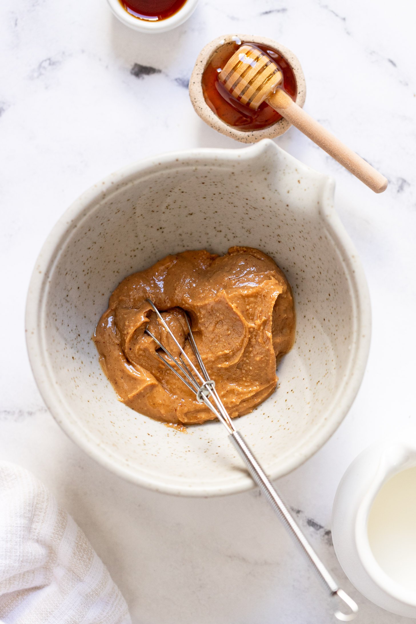 Mixing bowl with peanut butter and whisk next to honey.